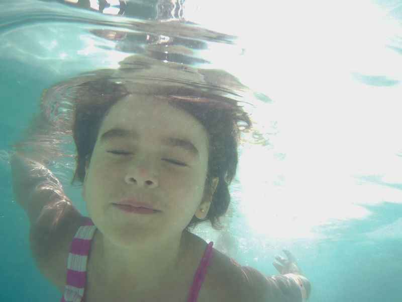 Underwater shot of a smiling girl swimming toward the camera, eyes closed, in sunlit turquoise water