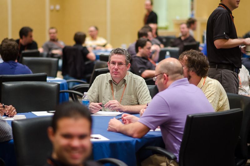 Conference attendees with lanyards seated at blue-draped round tables, ready for a workshop