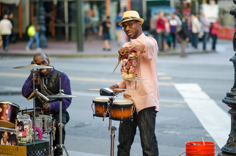 Two street musicians playing drums and bongos on a city sidewalk