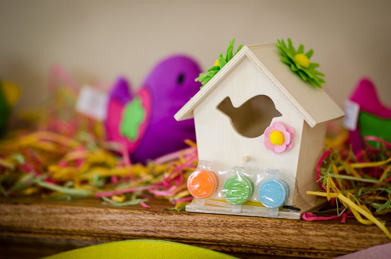 A small unpainted wooden birdhouse craft kit with paint pots, next to a purple plush bird