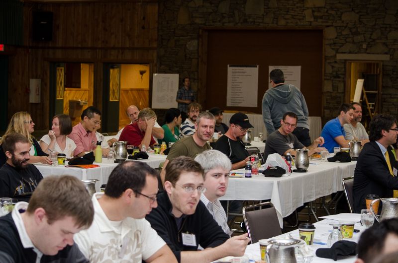 Conference attendees seated at long tables with coffee pots, waiting for a session to start