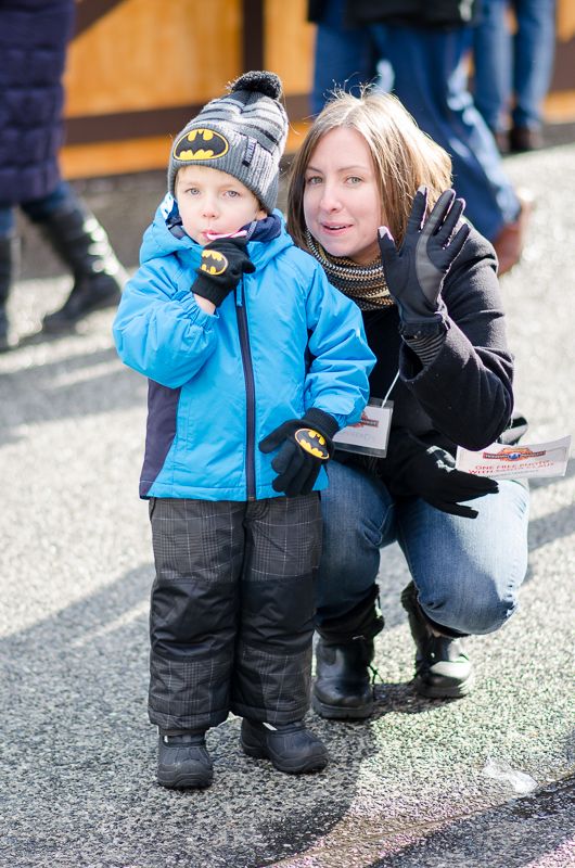 A boy in a blue snowsuit and Batman hat with a waving woman crouched beside him at an outdoor food drive