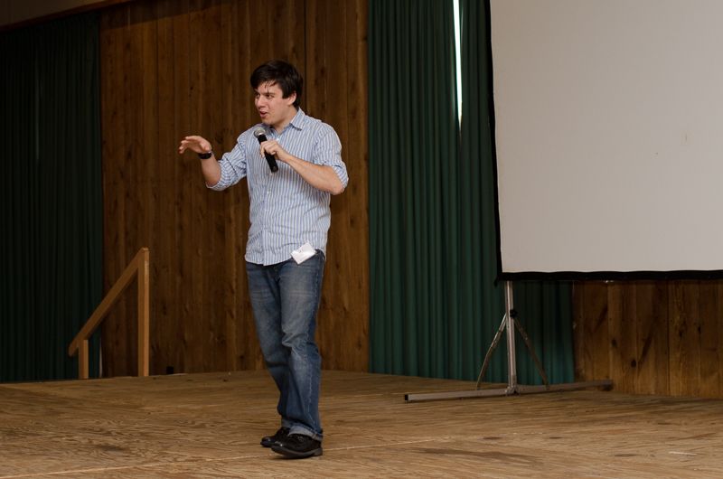A young speaker with a microphone gesturing on a wooden stage beside a projection screen