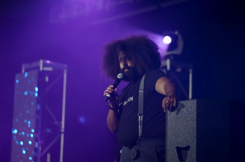 Comedian and musician Reggie Watts performing with a microphone under purple stage lights