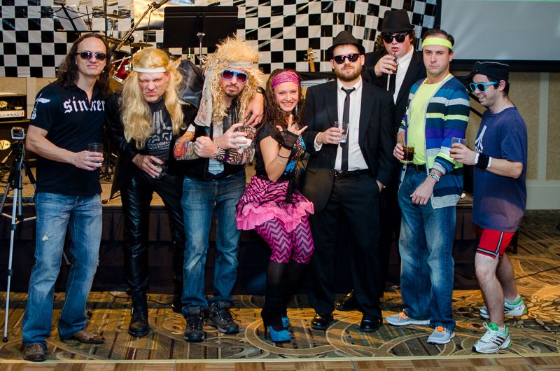 A group in 1980s costumes, from glam-rock wigs to neon headbands and Blues Brothers suits, posing on stage