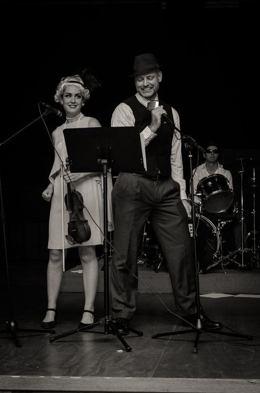 Black-and-white photo of a violinist and a singer in a fedora performing on stage in vintage outfits