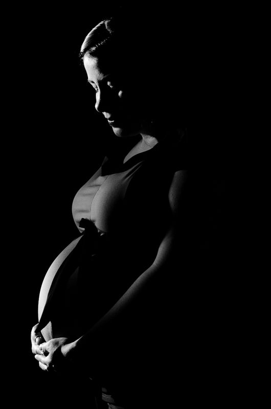 Low-key maternity portrait of a woman in silhouette, looking down with her hands around her belly