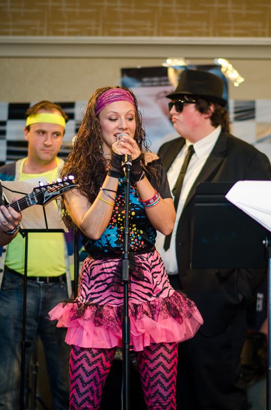 A singer in neon 80s style with a pink tutu and headband singing at the microphone with her band