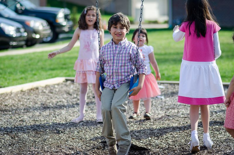 A grinning boy on a playground swing while girls in party dresses play around him