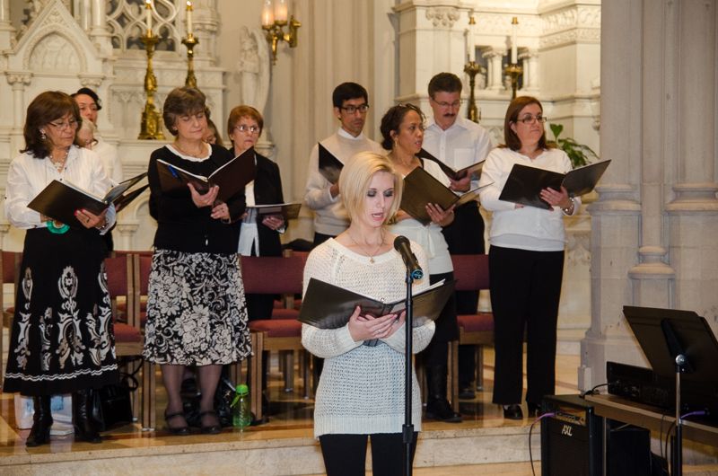 A church choir in black and white holding music folders while a young woman sings at the microphone