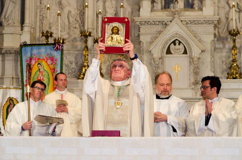 A bishop raising an ornate Gospel book at a marble altar, flanked by priests and a Virgin of Guadalupe banner