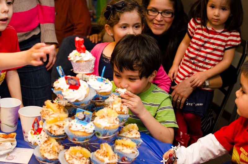 A boy taking a bite from a tower of cupcakes as other children and a mother crowd around the party table