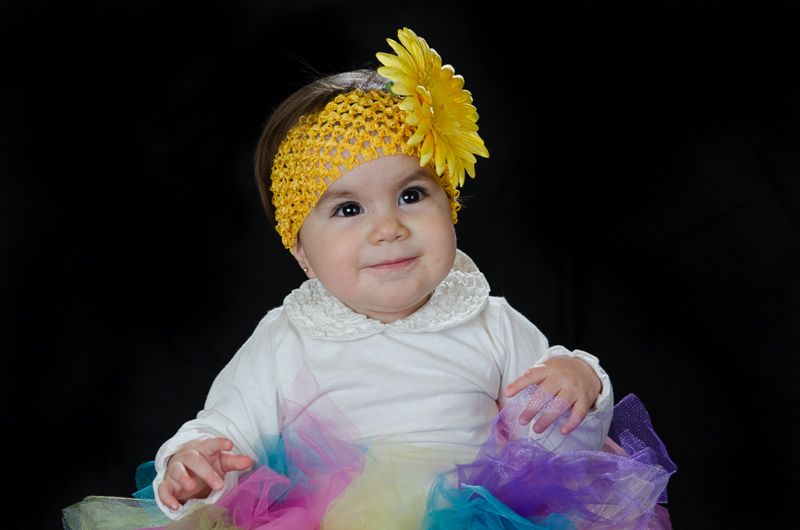 A smiling baby in a yellow sunflower headband and a rainbow tutu against a black backdrop