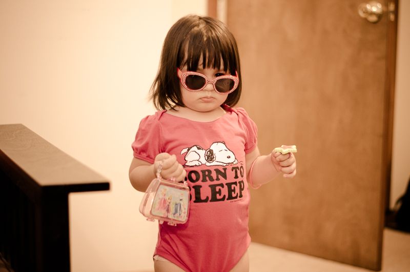 A toddler in pink heart-shaped sunglasses and a Snoopy onesie holding a tiny purse