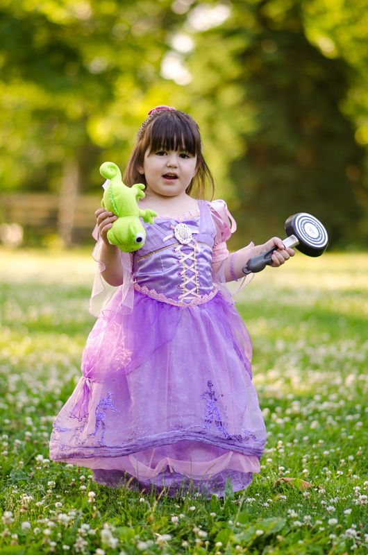 A little girl in a purple Rapunzel gown holding a toy frying pan and a green chameleon plush on the lawn