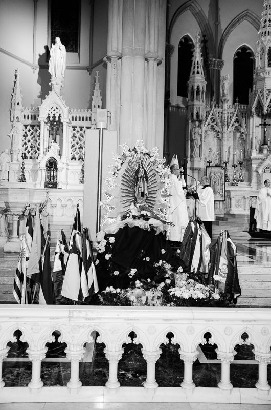 Black-and-white view of a Gothic altar where flags and flowers surround an image of the Virgin of Guadalupe