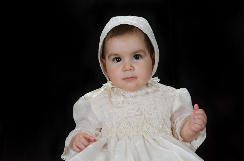 A baby in a white christening gown and bonnet against a black backdrop