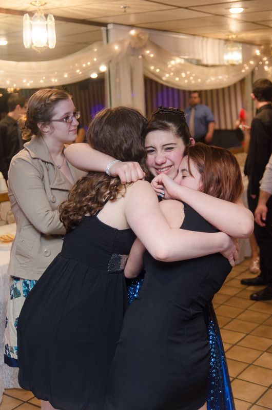 Three teenage girls in party dresses sharing a tight hug in a decorated ballroom