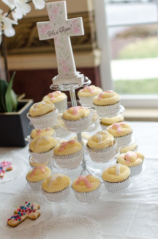 A tiered stand of cupcakes topped with pink fondant crosses beneath a decorative "He is Risen" cross
