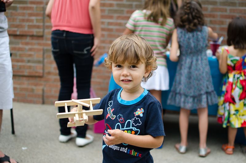 A curly-haired toddler smiling and holding a wooden toy airplane at an outdoor party