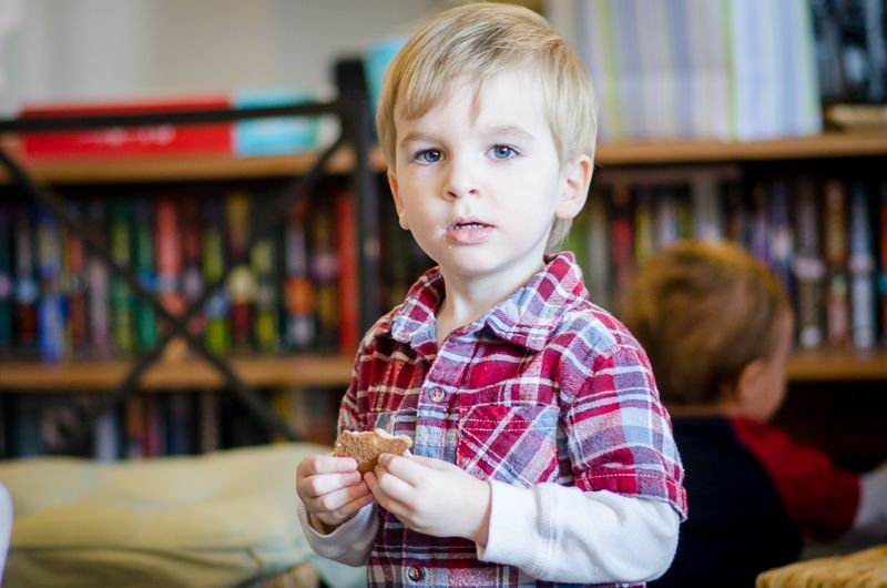 A blond boy in a plaid shirt holding a cookie in front of a bookshelf