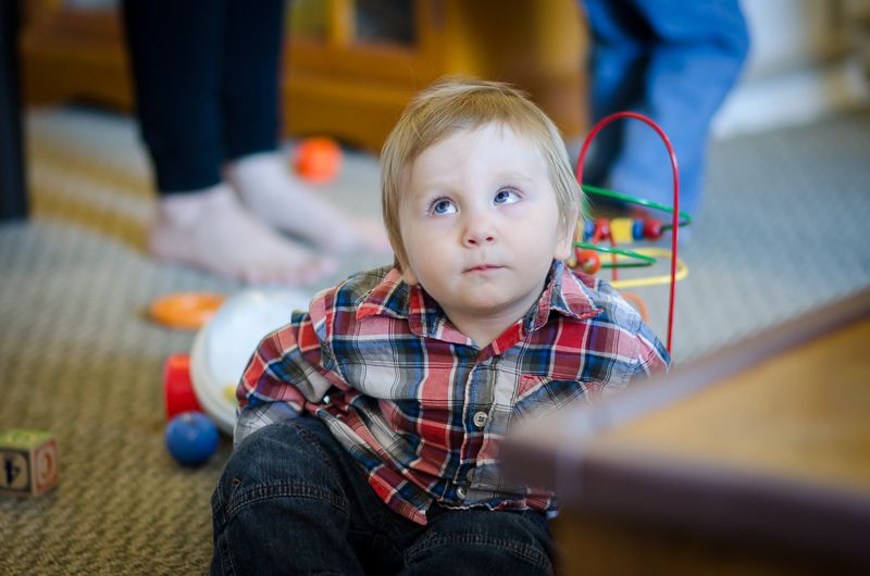 A blond toddler in a plaid shirt sitting on the carpet among toys, looking up