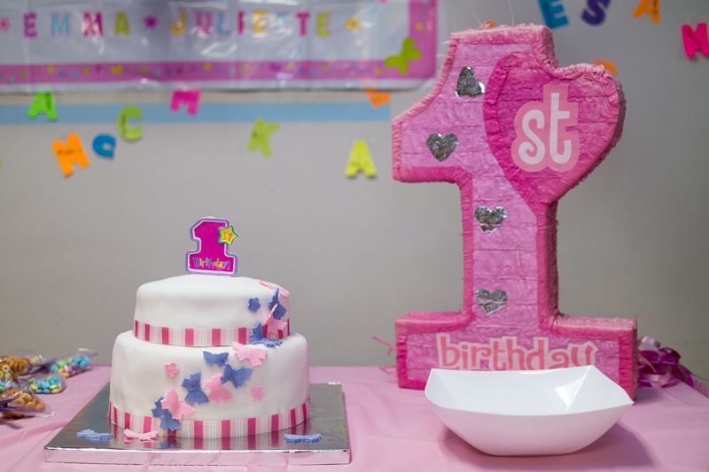 A pink and white two-tier first-birthday cake beside a pink "1st birthday" piñata