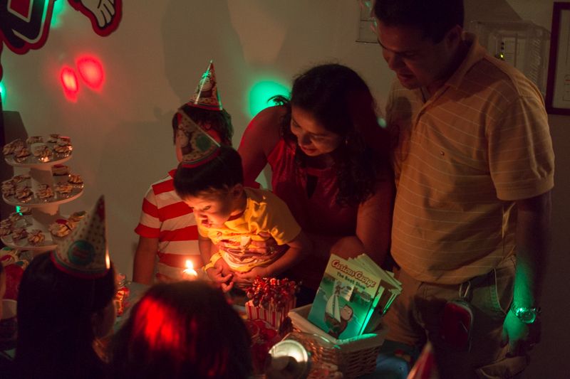 A boy in a party hat blowing out a birthday candle while his parents lean in, lit by the candle's glow