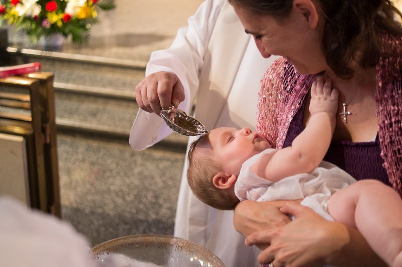 A priest pouring baptismal water over a baby's head as the godmother holds the baby over the font