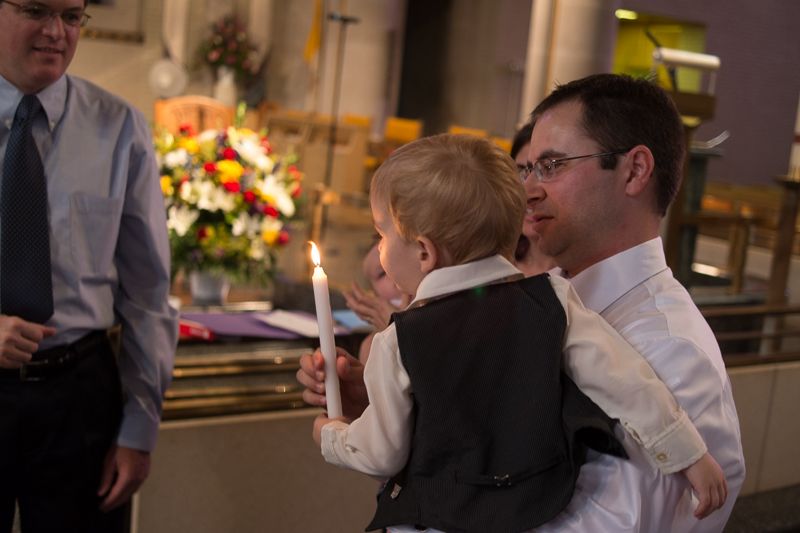 A toddler in a vest holding a lit baptismal candle in his father's arms