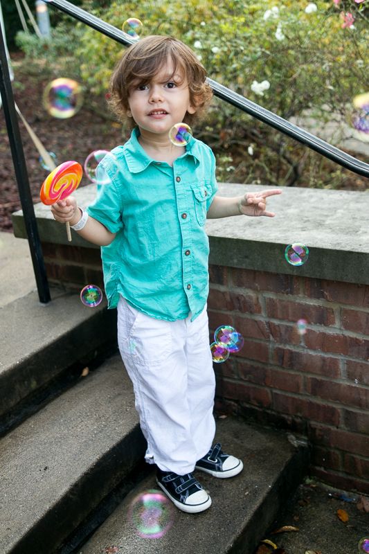 A curly-haired boy with a swirl lollipop standing on brick steps as soap bubbles float around him