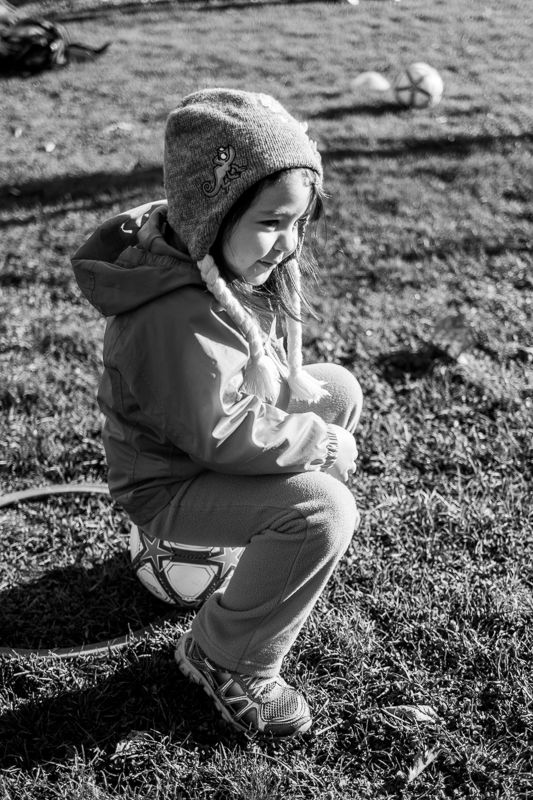 Black-and-white photo of a girl in a knit hat crouching on a soccer ball in the grass
