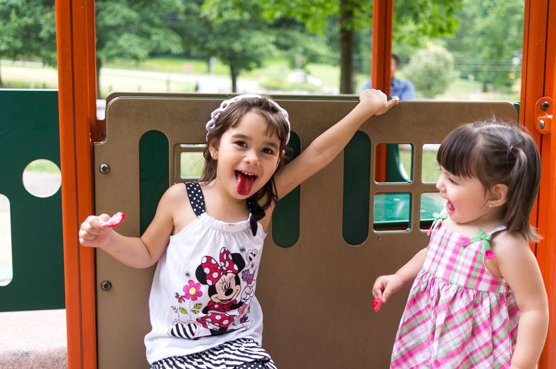 Two girls on a playground, one sticking out a tongue stained red by a lollipop