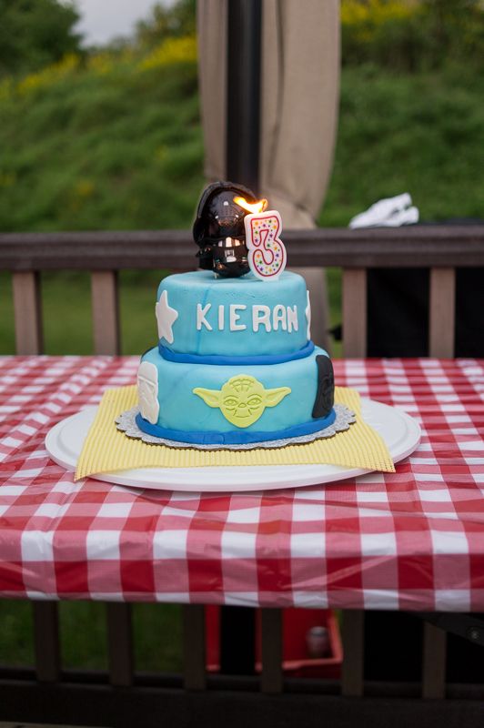 A two-tier blue Star Wars birthday cake with Yoda and Darth Vader on a red gingham tablecloth outdoors
