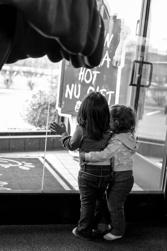 Black-and-white photo of two small children hugging as they look out a glass shop door
